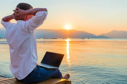 A man sitting with a laptop on a jetty at sunset, hands behind his head.