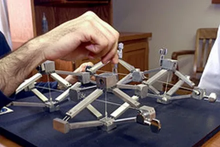 A hand pressing down on a model bridge built from interlocking metal struts.