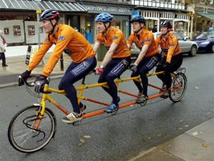 Four cyclists in matching orange kit riding a single long tandem bicycle past shops.